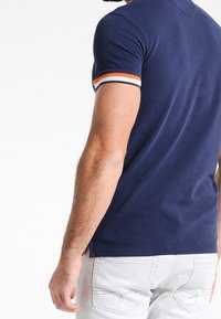 Námořnická modrá polokošile s krátkým rukávem, která má na rukávech oranžové a bílé pruhy, vyrobená z měkké a hladké tkaniny.