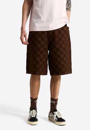 Personne portant un short à carreaux marron, un t-shirt blanc, des chaussettes marron Vans et des baskets Vans à carreaux noir et blanc, debout devant un fond uni.
