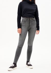 Osoba nosící šedé vysoké úzké džíny, černou krátkou mikinu, černé ponožky a černo-bílé tenisky na šněrování na bílém pozadí.