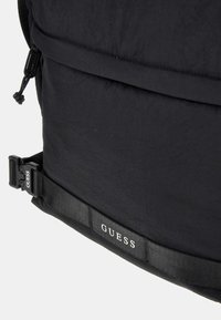 Rucsac negru cu țesătură texturată, curele ajustabile și o cataramă laterală. Prezintă un logo proeminent "GUESS" pe curea.