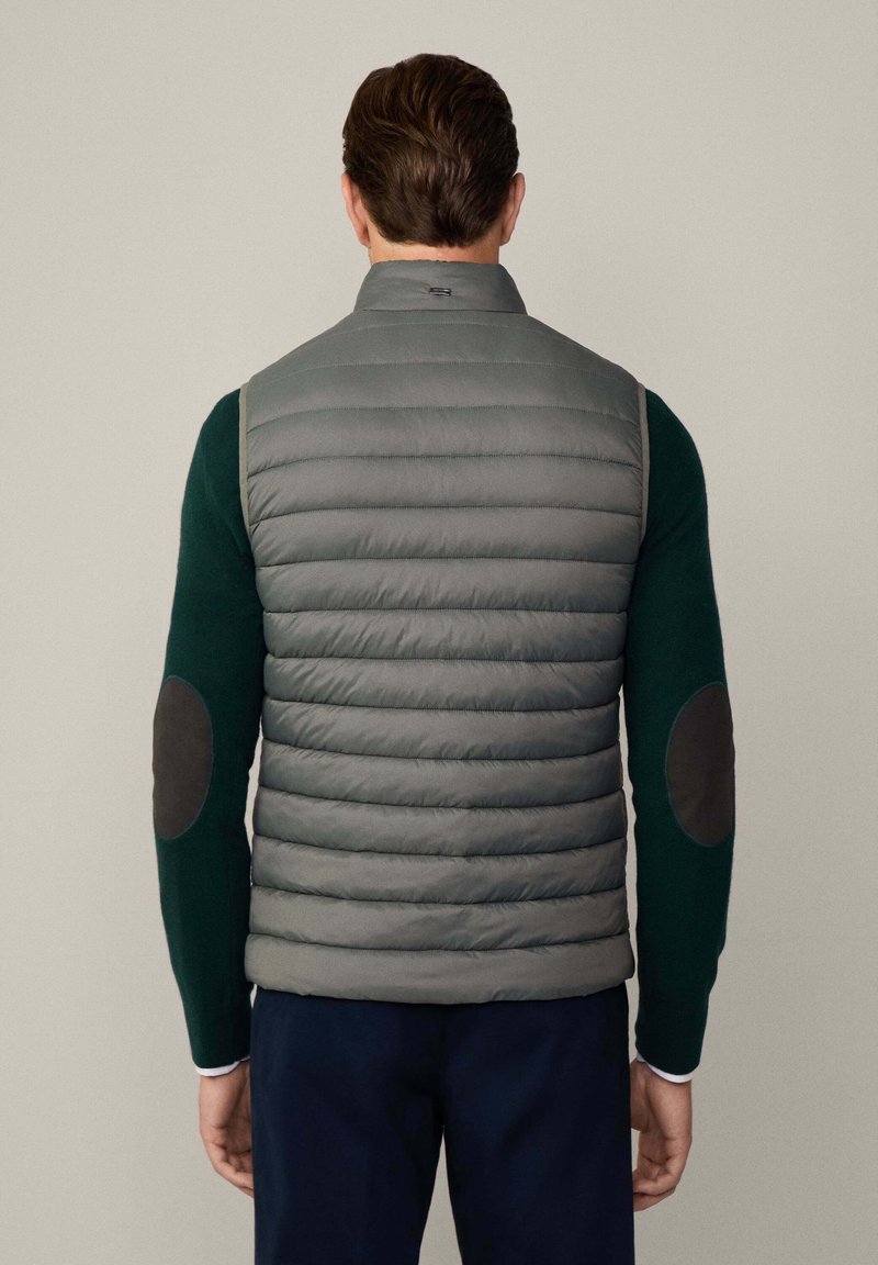 Grau gefütterte Weste mit hohem Kragen und horizontalem Steppdesign, getragen über einem grünen Pullover mit dunklen Ellbogenflicken, von hinten betrachtet.