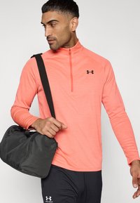 Ελαφρύ, μακρυμάνικο τοπ με υφή σε κοραλί χρώμα, με γιακά μισού φερμουάρ και μαύρο λογότυπο· συνδυασμένο με σκούρο παντελόνι και μαύρη τσάντα τύπου duffel.