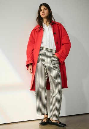 Woman standing with hand in striped pants pocket, wearing a red coat, white shirt, and black ballet flats against a plain light wall.