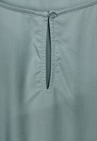 Tessuto della blusa di colore verde chiaro con scollatura rotonda, caratterizzata da una piccola apertura fissata da un bottone al centro. Texture liscia, senza motivi.