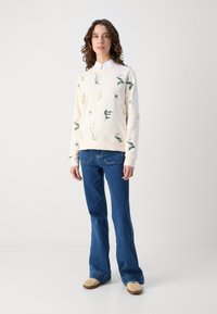 Langermet kremfarget genser med blomsterbroderi, kombinert med blå flagrende jeans og beige fottøy. Tekstilt, avslappet antrekk med botaniske detaljer.