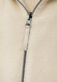 Nahaufnahme einer cremefarbenen, strukturierten Fleecejacke mit einem glanzsilbernen Reißverschluss und einem Stoffziehband, das mit "S. Oliver" beschriftet ist.