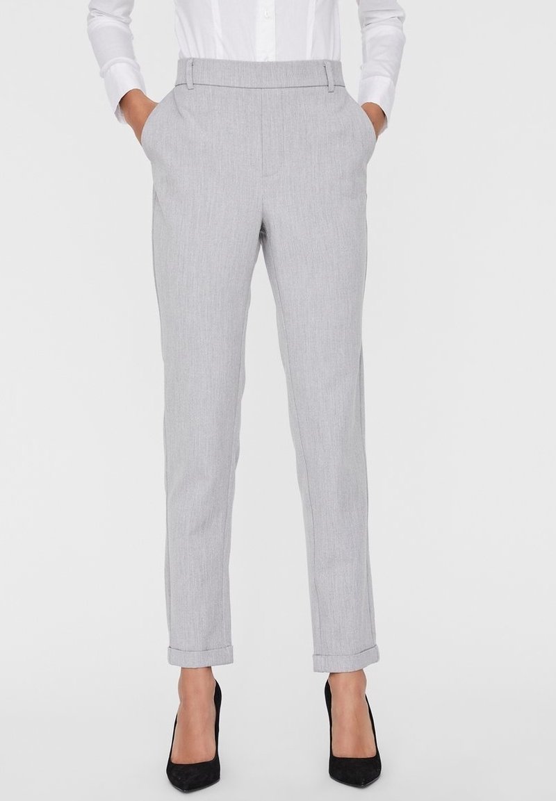 Femme portant un pantalon gris clair cintré avec des revers, mains dans les poches, chemise blanche et escarpins noirs pointus, se tenant sur un fond uni.