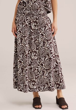 Žena oblečená v čierno-bielej maxi sukni s mierne nariaseným pásom a čiernych otvorených sandáloch na podpätku, stojaca pred jednoliatym pozadím.