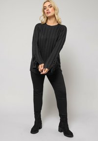Czarny sweter prążkowany z długimi rękawami, ozdobiony detalami w postaci kokardek przy mankietach, zestawiony z czarnymi dopasowanymi spodniami i botkami na obcasie.