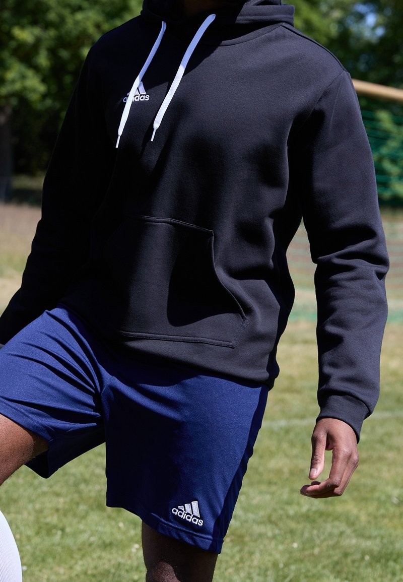 Sudadera negra con cordones blancos y un bolsillo delantero, combinada con pantalones cortos azules que tienen un logo blanco en el dobladillo. Fondo de campo de hierba.