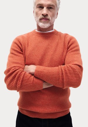 Orange knit sweater with textured pattern, round neckline, and ribbed cuffs. Worn over dark jeans, arms crossed.