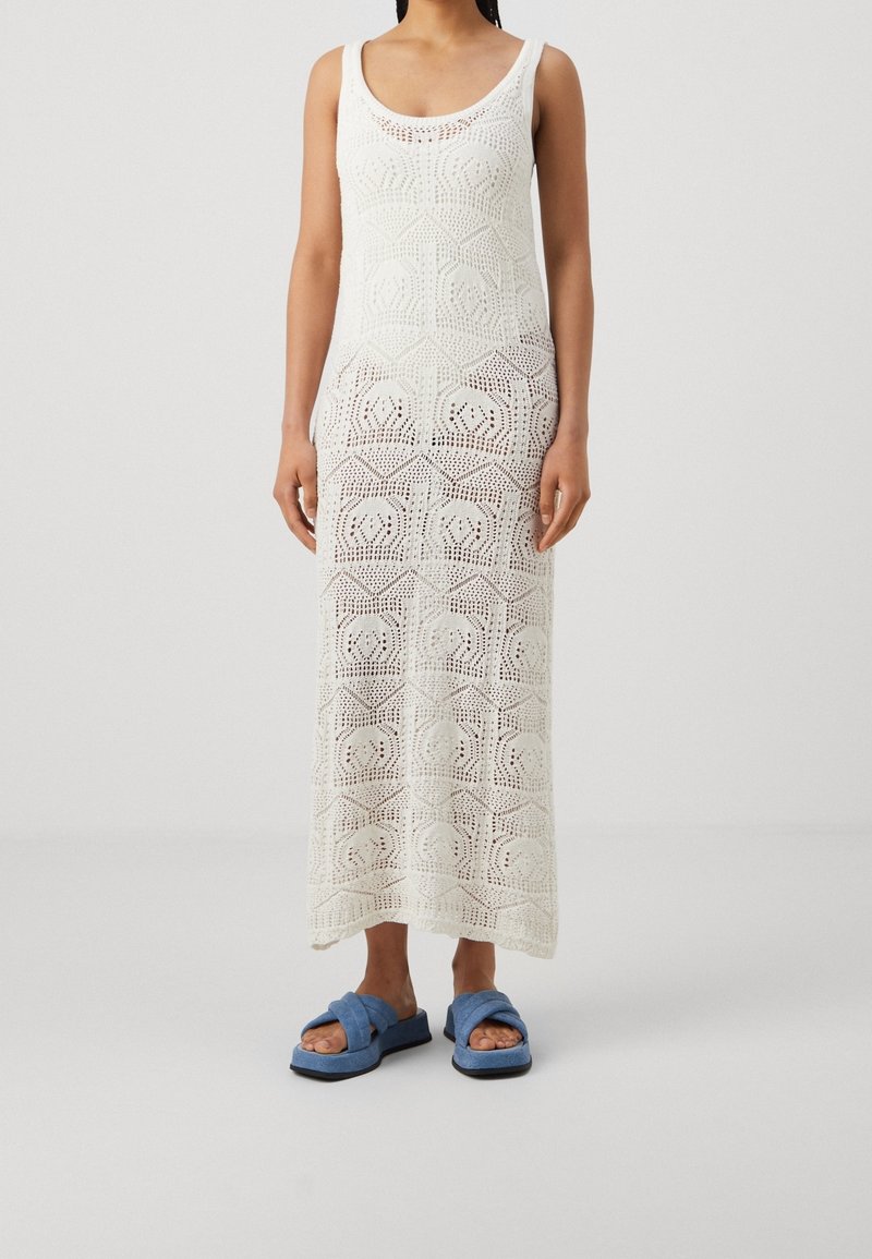 Femme portant une robe maxi blanche en crochet sans manches et des sandales à glissière bleues rembourrées sur un fond clair uni.