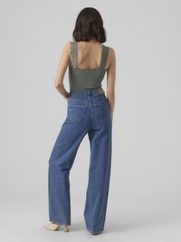 Vrouw in een groene top met kantafwerking, hoge taille wijde spijkerbroek en beige puntige hakken, staand met haar rug naar de kijker gekeerd.