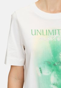 Weißes T-Shirt mit kurzen Ärmeln, das einen grünen Farbverlauf zeigt, mit dem Schriftzug "UNLIMITED" in grüner Glitzerfarbe und Verzierungen, die florale Muster hervorheben.