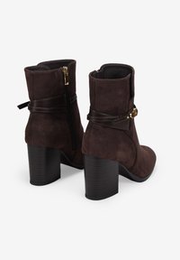 Botas de tobillo de ante marrón con un tacón macizo acanalado, cremallera lateral y una correa de cuero decorativa con un acento dorado.