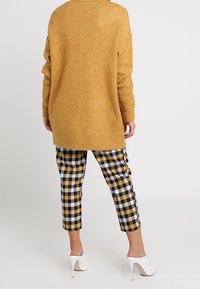 Sinappikeltainen villapaita, löysä istuvuus, pitkät hihat; yhdistetty mustavalko-keltaisiin ruudullisiin polvipitkiin housuihin; valkoiset korkokengät.