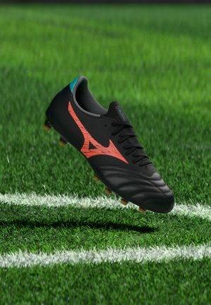 Čierny futbalový kopačka s červeným logom a žltými kolíkmi vznášajúci sa nad zelenou trávou blízko bielych čiar ihriska.