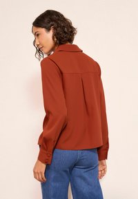 Rozsdaszínű blúz gallérral, hosszú ujjak gombdíszítéssel és hátsó redővel. Kék farmernadrággal párosítva.