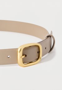 Ζώνη από μπεζ δέρμα με ομαλή υφή, με τετράγωνη χρυσή μεταλλική αγκράφα και πέντε τρύπες ρύθμισης για ευελιξία στο μέγεθος.