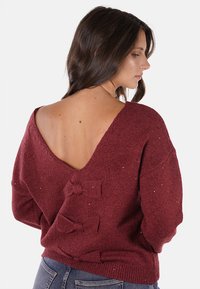 Maglione lavorato a maglia borgogna con un profondo scollo a V sul retro, caratterizzato da un dettaglio a fiocco decorativo di lato e una finitura texturizzata. Maniche lunghe, vestibilità comoda.