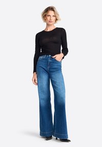Schwarzes Langarmoberteil, kombiniert mit weit geschnittenen blauen Jeans, die einen sanften Farbverlauf und eine subtile Textur aufweisen. Abgerundet mit schwarzen High Heels.
