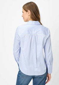 Camicia a righe celesti e bianche con colletto, maniche lunghe, polsini con bottoni e dettaglio arricciato sul retro. Tessuto di cotone liscio.