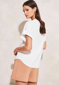 Top bianco dalla vestibilità ampia con maniche corte arrotolate, orlo curvo e texture liscia, abbinato a pantaloni corti beige. Design semplice senza motivi.