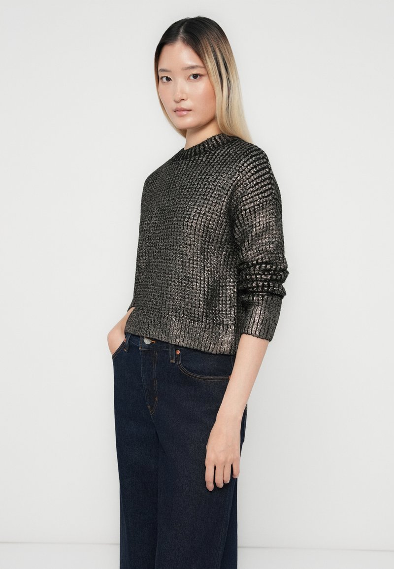 Donna con lunghi capelli biondi lisci che indossa un maglione lucido nero e argento e jeans blu scuri, in piedi con una mano in tasca.