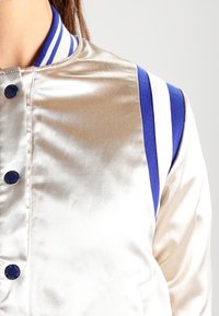 Giacca bomber in satin metallizzato di colore crema, con bordo a strisce blu e bianche e bottoni blu scuro.