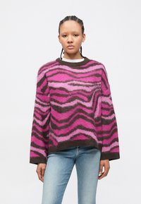 Jovem com cabelo trançado, vestindo um suéter felpudo rosa com riscas de zebra castanhas e jeans azul claro, contra um fundo branco.