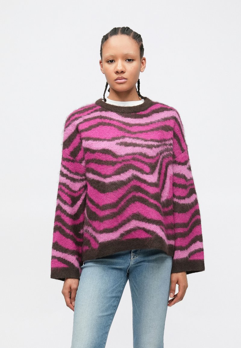 Jovem com cabelo trançado, vestindo um suéter felpudo rosa com riscas de zebra castanhas e jeans azul claro, contra um fundo branco.