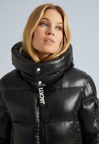 Blonde sieviete, kas valkā spīdīgu melnu pūkaino jaku ar augstu mīkstās daļas apkakli un JACKTIT logo uz priekšējā rāvējslēdzēja, pret vienkāršu fona.