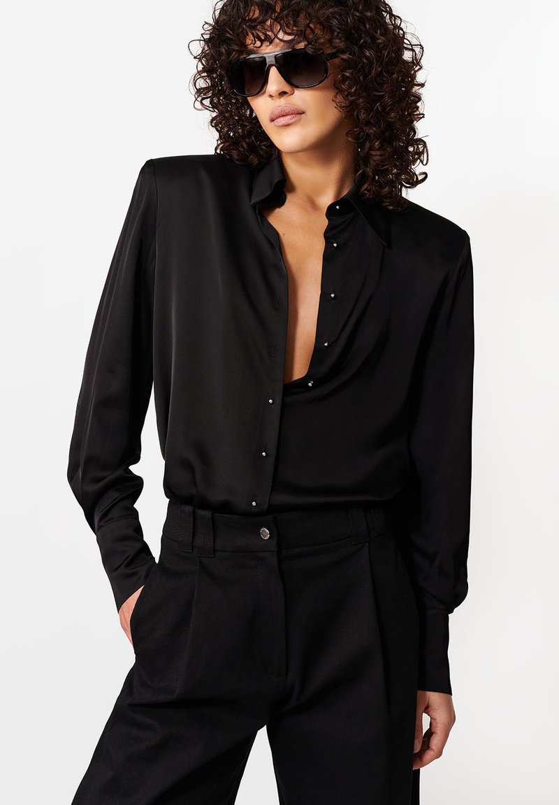 Osoba s kučeravými vlasmi, ktorá má na sebe čierne slnečné okuliare, voľnú čiernu saténovú košeľu so zapnutými gombíkmi, ktorá je čiastočne rozopnutá, a čierne vysoký pás nohavíc.