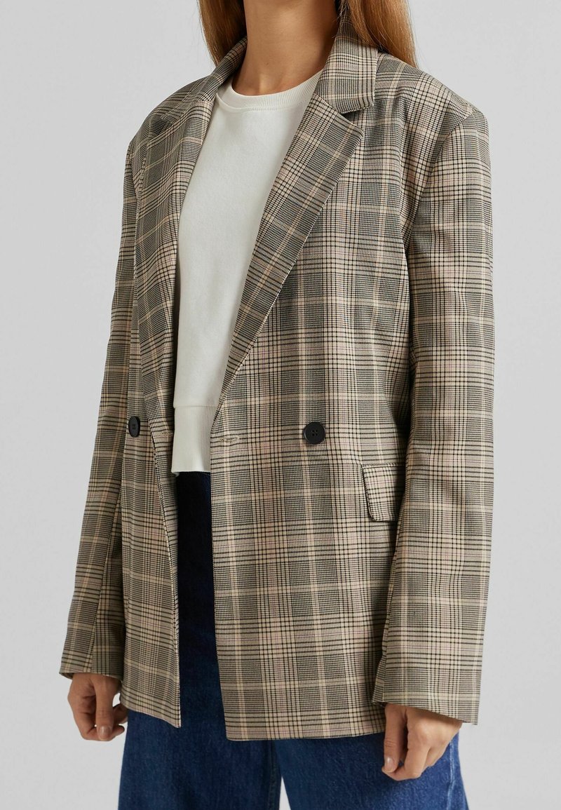 Blazer a cuadros en beige, marrón y negro con un corte relajado, solapas con muescas, doble botonadura y bolsillos laterales. Llevado sobre una camiseta blanca y pantalones azules.
