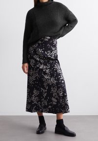 Pull en maille noire épaisse avec un col montant, associé à une jupe midi en satin noire à motif floral avec des motifs de feuillage gris. Mocassins noirs et chaussettes.