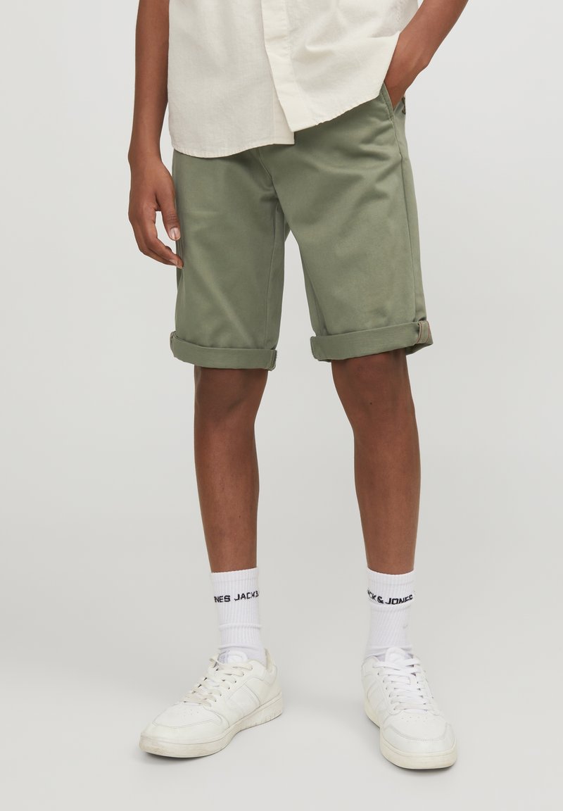 Personne portant un short olive retroussé, des chaussettes blanches avec un texte noir, des baskets blanches et une chemise beige avec une main dans la poche.