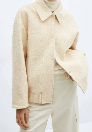 Personne portant une veste en laine beige sur un pull à col roulé clair avec un pantalon couleur crème, main reposant près de la poche.