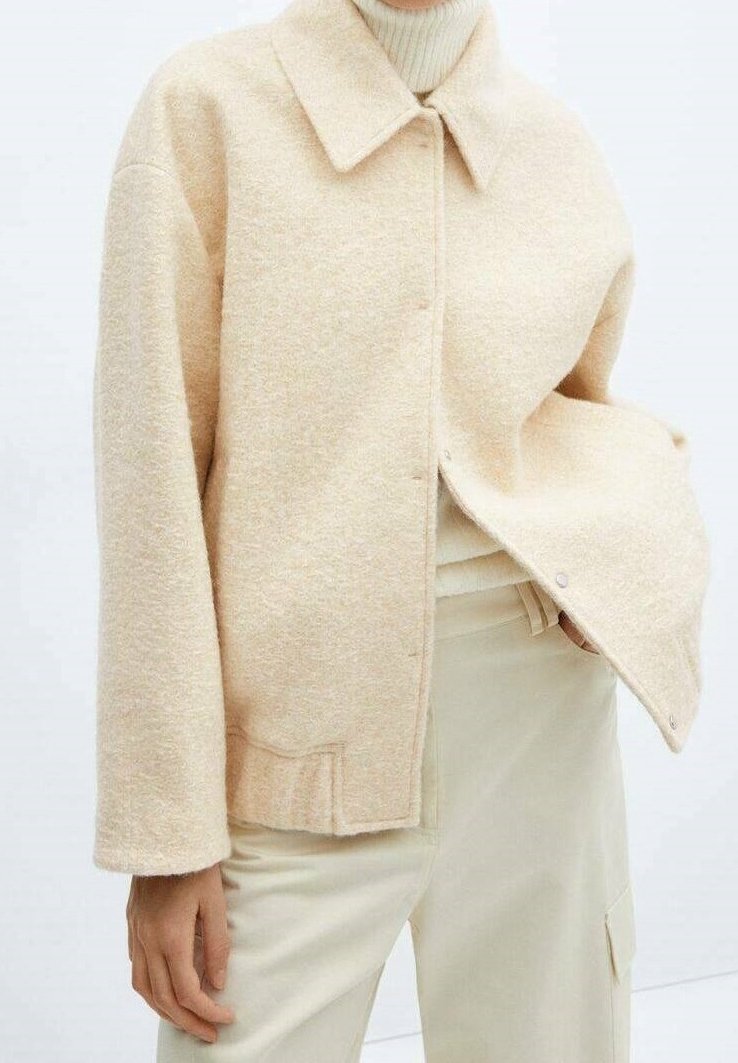 Personne portant une veste en laine beige sur un pull à col roulé clair avec un pantalon couleur crème, main reposant près de la poche.