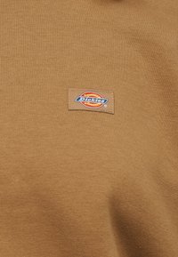 Καφέ ύφασμα με μικρή ορθογώνια ετικέτα όπου φαίνεται το λογότυπο Dickies σε κόκκινο, κίτρινο, μπλε και λευκό χρώμα.