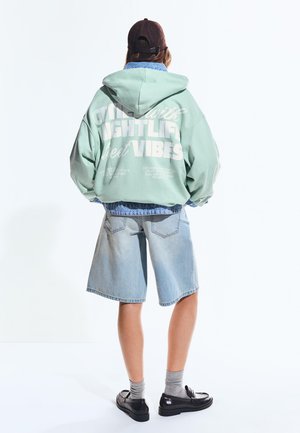 Személy menta zöld kapucnis pulóverben, fehér felirattal, farmer rövidnadrágban, szürke zokniban, fekete cipőben és sötétbarna sapkában, háttal állva.