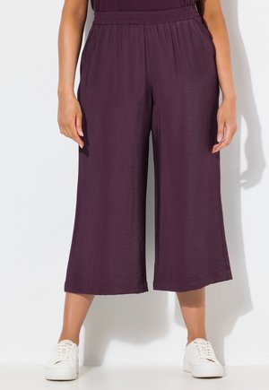 Femme portant un pantalon ample et court violet foncé avec une ceinture élastique et des baskets blanches à lacets, debout devant un fond blanc uni.