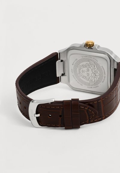 Fond carré en argent de montre avec gravure de Méduse, bracelet en cuir marron et boucle en argent gravée du nom de la marque.