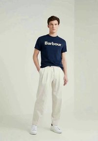 Νεαρός άνδρας στέκεται φορώντας ένα ναυτικό μπλε t-shirt Barbour, κρεμ παντελόνια με πτυχώσεις και λευκά sneakers μπροστά σε ένα απλό ανοιχτό φόντο.