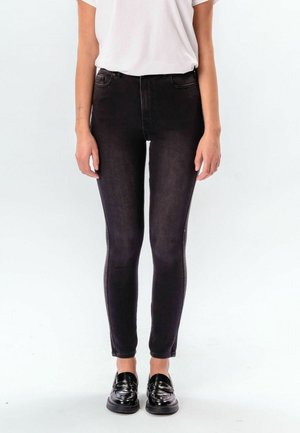 Nő szűk fekete farmerben, fehér rövid ujjú ingben és fekete mokaszinban, sima fehér háttér előtt állva.