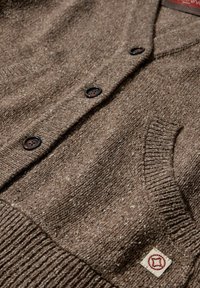 Brauner Cardigan aus strukturiertem Strickstoff, mit V-Ausschnitt, drei dunklen Knöpfen und einem Patch mit einem roten Emblem auf der Tasche.