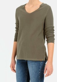 Oliwkowy sweter w serek z długimi rękawami, teksturowana dzianina, boczne rozcięcia oraz małe logo na dole. Noszony na niebieskich dżinsach.