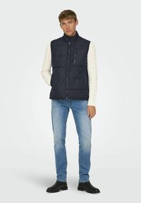 Jeune homme portant un gilet matelassé noir par-dessus un pull en mailles blanches, un jean bleu clair et des bottes noires, se tenant avec une main dans sa poche.