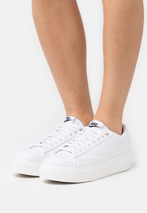 Zapatillas de lona blancas con un acabado texturizado, suela de goma blanca plana y logo azul marino en la lengüeta, detalladas con ojales dorados.