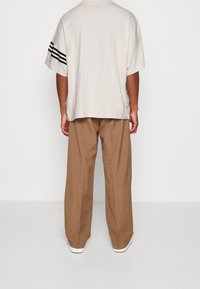 Ljusbeige oversized t-shirt med svarta ränder på ärmarna, tillsammans med löst sittande bruna byxor, sedd bakifrån.