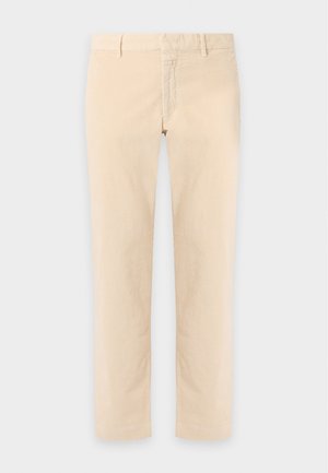 Homme debout les mains dans les poches, portant une chemise blanche à manches courtes avec col, un pantalon beige, une ceinture noire, et des mocassins noirs et blancs.